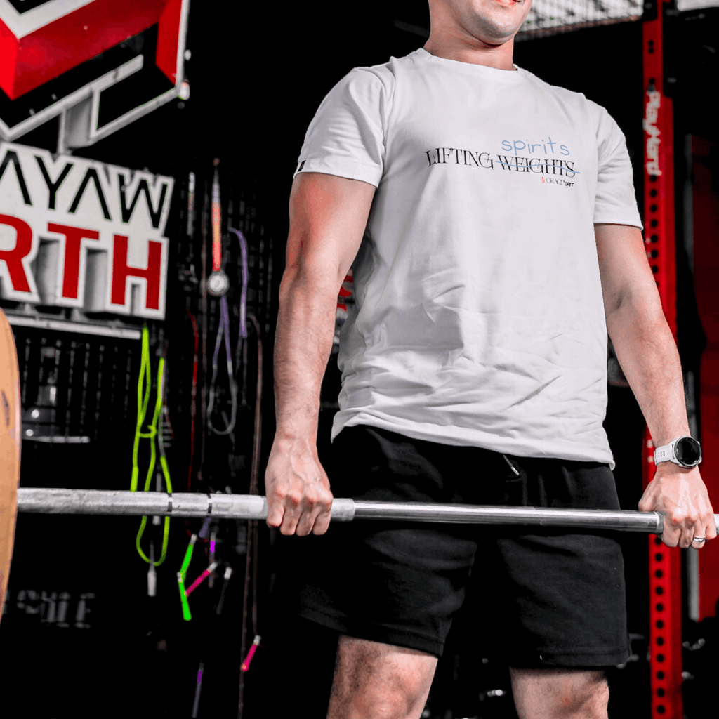 Man lifting a barbell in gym, wearing a white t-shirt with a print that says 'LIFTING spirits 𝖶̶𝖤̶𝖨̶𝖦̶𝖧̶𝖳̶𝖲̶' 
