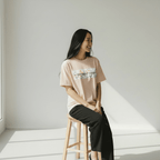 Woman sitting on a stool wearing a peach t-shirt with text and graphics, in a bright room.