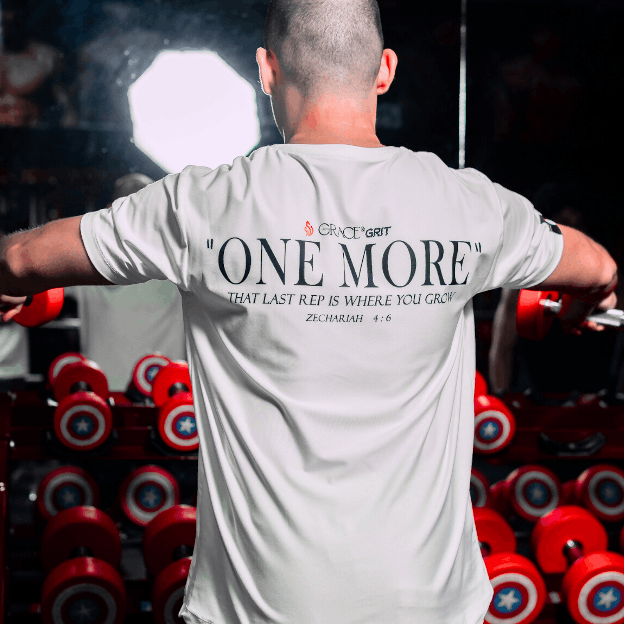 A man lifting weights with a white shirt on displaying motivational text with Zechariah 4:6, in a gym setting