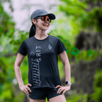 Woman wearing a mint and black running shirt with green and white text, standing outdoors with greenery in the background