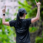 A woman facing away wearing a mint and black running shirte text, standing outdoors with arms raised pointing to the sky, greenery in the background.