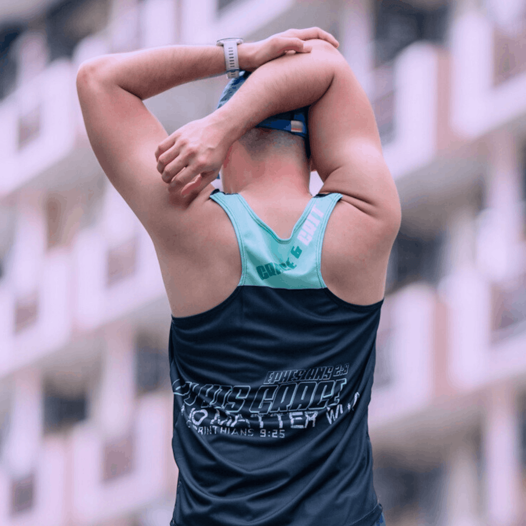A gentleman stretching outdoors wearing a mint and black singlet, facing away from camera and wearing a cap.