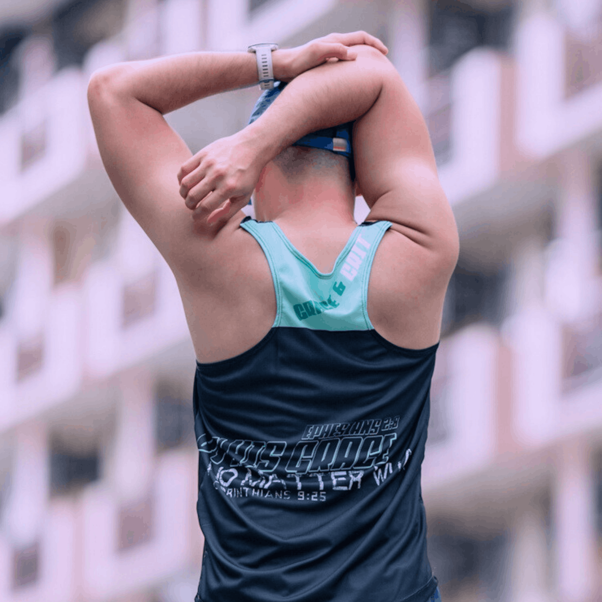 A gentleman stretching outdoors wearing a mint and black singlet, facing away from camera and wearing a cap.
