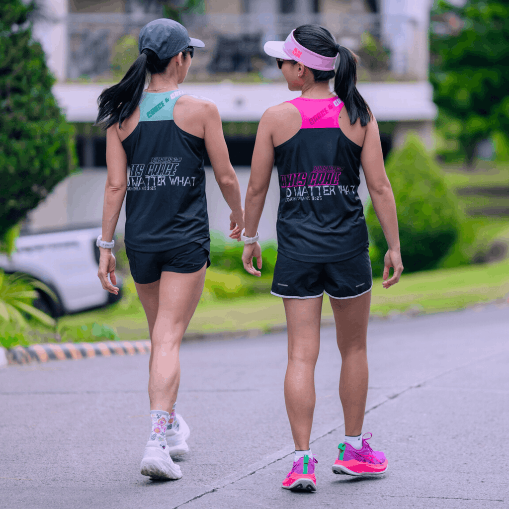 Two women walking outdoors wearing the mint and black and the other one wearing the pink and black running singlet.