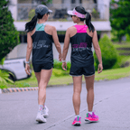 Two women walking outdoors wearing the mint and black and the other one wearing the pink and black running singlet.