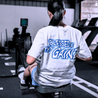 A woman working out in the gym wearing a white t-with a print that says 'BLESSED BE THY GAINZ' and "'GOD'S STRENGTH UNTO YOU' PHILIPPIANS 4:13".