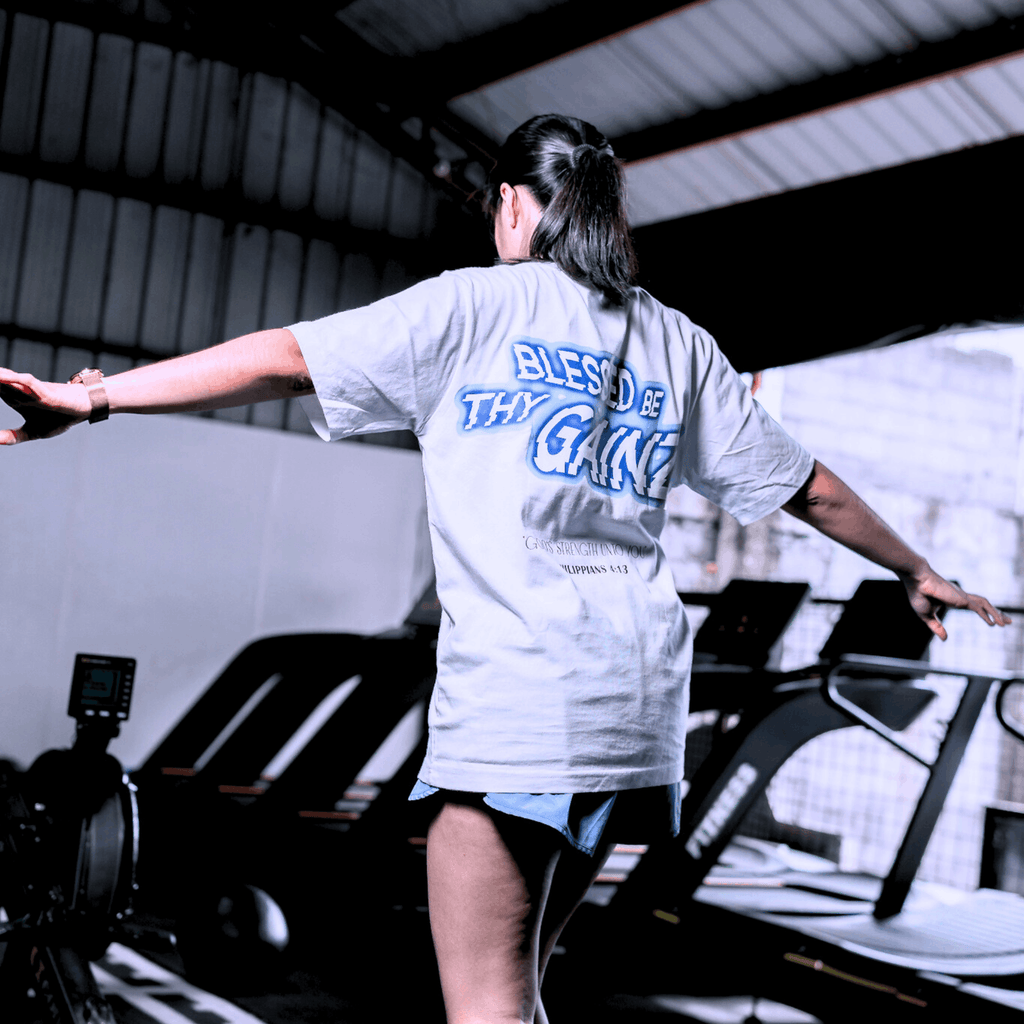 A woman in the gym wearing a white t-with a print that says 'BLESSED BE THY GAINZ' and "'GOD'S STRENGTH UNTO YOU' PHILIPPIANS 4:13".