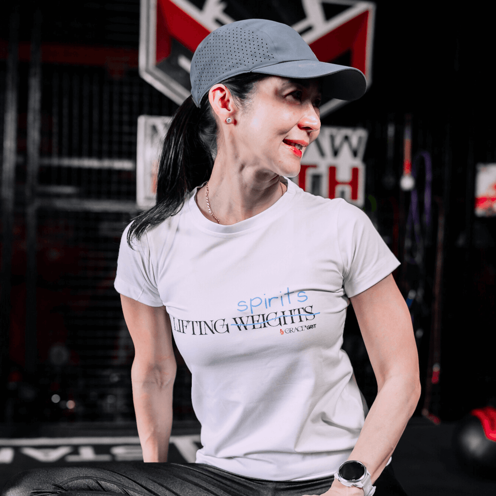 A woman in the gym wearing a white t-shirt with a print that says 'LIFTING spirits 𝖶̶𝖤̶𝖨̶𝖦̶𝖧̶𝖳̶𝖲̶' 